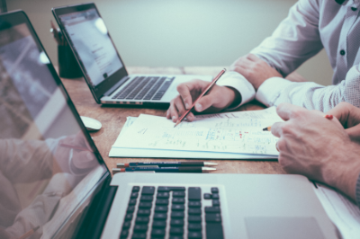 Two people working together, photo of two laptops, one hand writing on paper between them. Photo by Scott Graham on Unsplash. Help people!